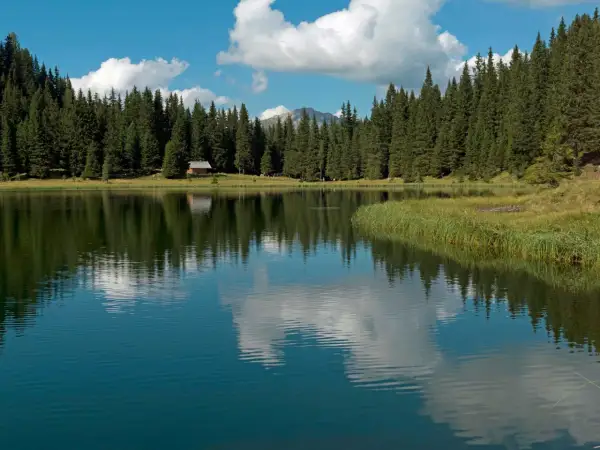 Riflessi di un cielo azzurro e nuvoloso sul lago di Cece, circondato da fitti abeti e una piccola baita.