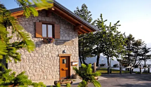 Rifugio Predaia, un'accogliente costruzione in pietra con tetto spiovente, circondato da alberi verdi e un cielo azzurro.