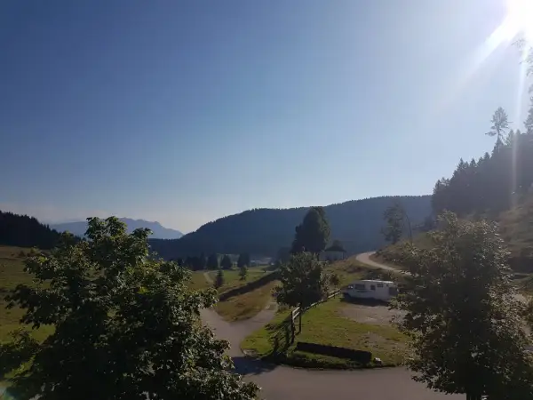 Un camper parcheggiato vicino a un **rifugio barricata** in montagna, con un sentiero che si snoda tra prati verdi e boschi.