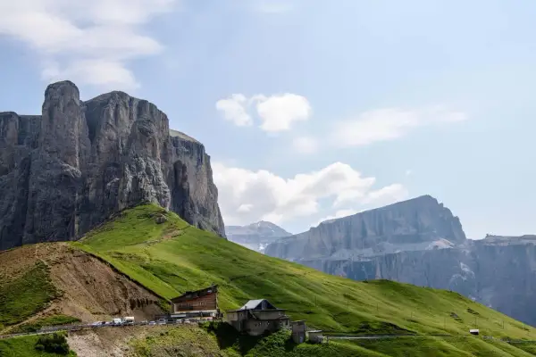Edifici alpini e un parcheggio affollato ai piedi di imponenti pareti rocciose sul passo Sella, con verdi pendii erbosi.