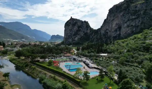 Agricamp Altogarda: un villaggio vacanze con piscine e scivoli ai piedi di imponenti montagne, vicino a un fiume e un borgo.