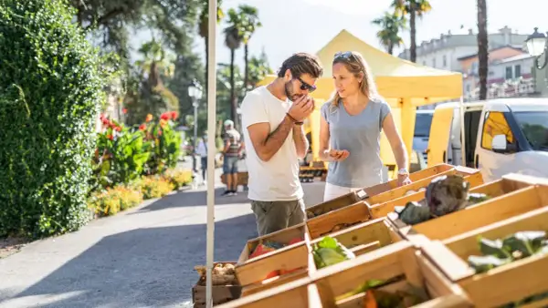 Coppia al markt Riva del Garda, annusando prodotti freschi. Verdure in cassette di legno sotto il sole.