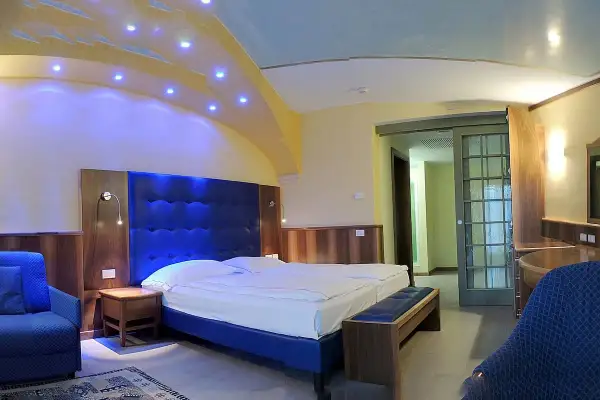 Camera moderna con letto king size e testiera blu imbottita, illuminazione soffusa e soffitto decorato. Un assaggio del comfort del Grand Hotel Astoria Lavarone.