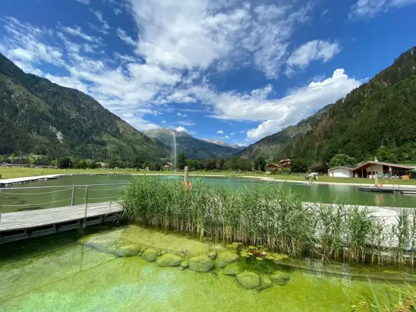 Un rinfrescante biolago a Predazzo, circondato da montagne verdi e un cielo azzurro con nuvole bianche. Pontili in legno e canneti creano un'atmosfera serena.