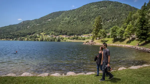 Coppia passeggia sul lungolago del lago delle piazze, con montagne verdi e un villaggio sullo sfondo.