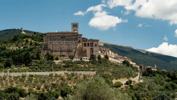 La Basilica di San Francesco domina il colle di Assisi, circondata da uliveti e montagne verdi sotto un cielo azzurro con nuvole bianche.