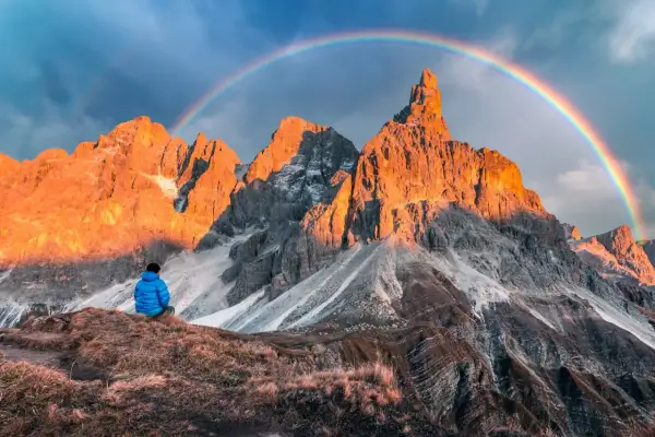 Persona seduta ammira le maestose Pale di San Martino illuminate dal sole al tramonto, con un arcobaleno che le sovrasta.