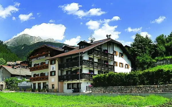 Albergo La Montanara a Predazzo, con le sue balconate in legno e le montagne sullo sfondo, incorniciato da un cielo azzurro e nuvole bianche.