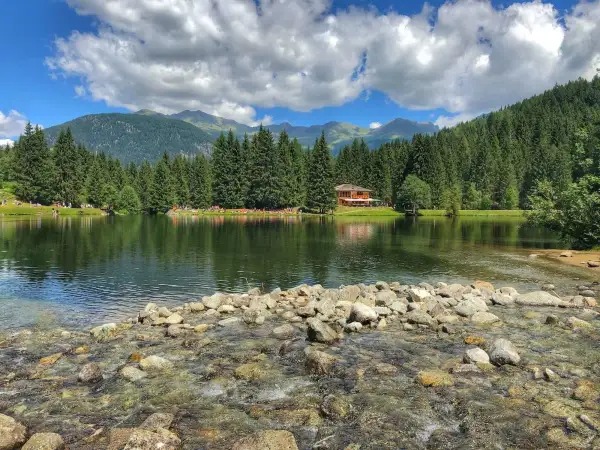 Il lago dei caprioli, circondato da fitti boschi e montagne, riflette un cielo azzurro con nuvole bianche. Una casa in legno si affaccia sull'acqua.