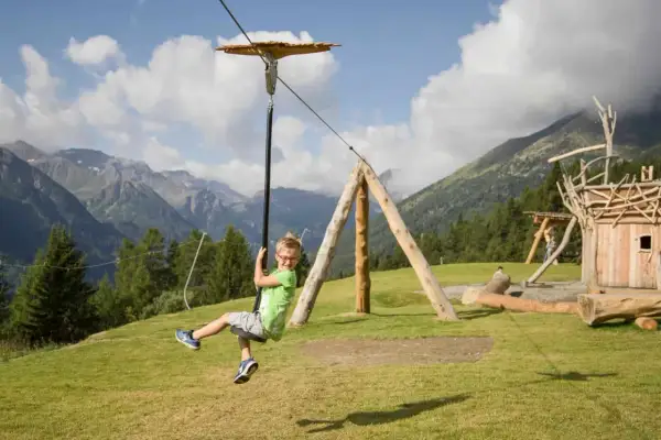 Bambino felice sfreccia su una teleferica nel **Pejo Kinderland**, con montagne sullo sfondo.