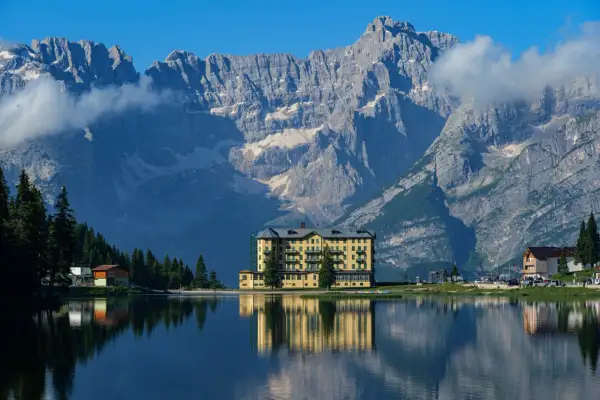 Un hotel giallo si specchia in un lago alpino, con imponenti montagne sullo sfondo: ecco dove si trovano le Dolomiti.