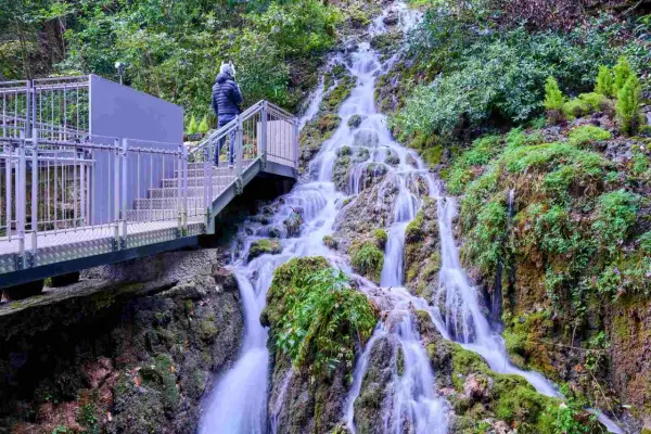 Una persona osserva le **cascate del Varone** da una passerella metallica. L'acqua scende a cascata su rocce ricoperte di muschio, immersa nel verde.