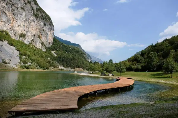 Un pontile in legno serpeggia sul lago di Andalo, circondato da montagne verdi e un cielo azzurro con nuvole bianche.