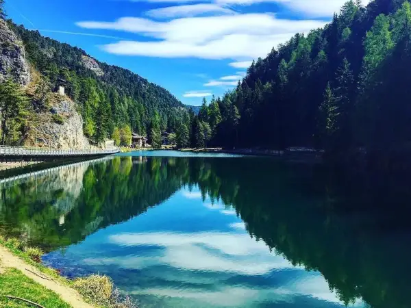 Un lago smeraldo incastonato tra montagne boscose. Una spiaggia erbosa invita al relax, mentre un ponte attraversa le acque calme.