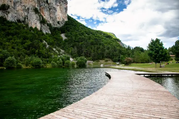 Un pontile in legno curva dolcemente sul lago di Nembia, circondato da montagne verdi e un cielo nuvoloso.