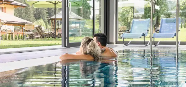 Coppia rilassata nella piscina interna di un wellness hotel Dolomia, con vista sul giardino e sulle montagne.