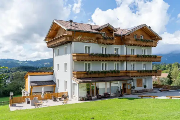 L'albergo Miramonti, con le sue balconate fiorite e il tetto in legno, si staglia contro un cielo azzurro con nuvole bianche, immerso in un paesaggio montano verdeggiante.