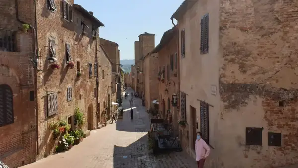 Una persona con mascherina passeggia per una stretta via acciottolata di **Certaldo Alto**, circondata da antichi edifici in mattoni con persiane in legno.