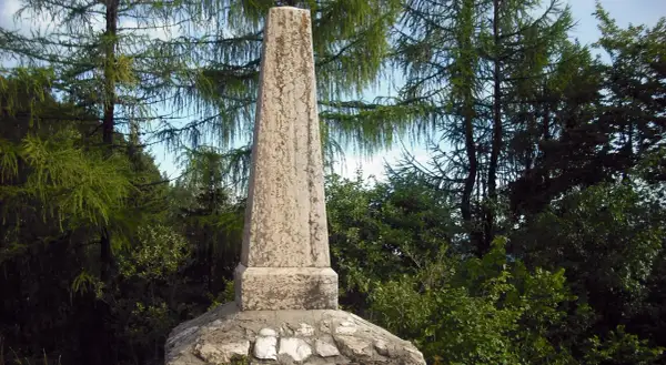 Obelisco in pietra sul **monte Zugna**, circondato da alberi di larice e vegetazione rigogliosa.
