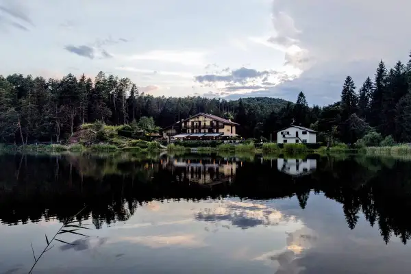 Riflessi serali sul lago di Santa Colomba, con un albergo e una casa immersi nel verde degli alberi.